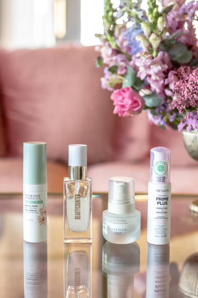 Four makeup primers lined up on a glass table with flowers in the background.