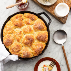 Top down view of biscuits in a cast iron skillet, condiments on the table, a wooden cutting board, and serving utensils on a gray and white marble surface.