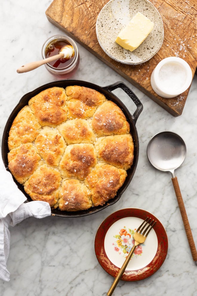 Top down view of biscuits in a cast iron skillet, condiments on the table, a wooden cutting board, and serving utensils on a gray and white marble surface. 