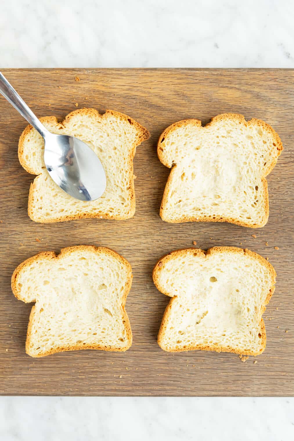 Spoon pressing down on a slice of bread sitting on a wooden cutting board with 4 other pieces of bread.