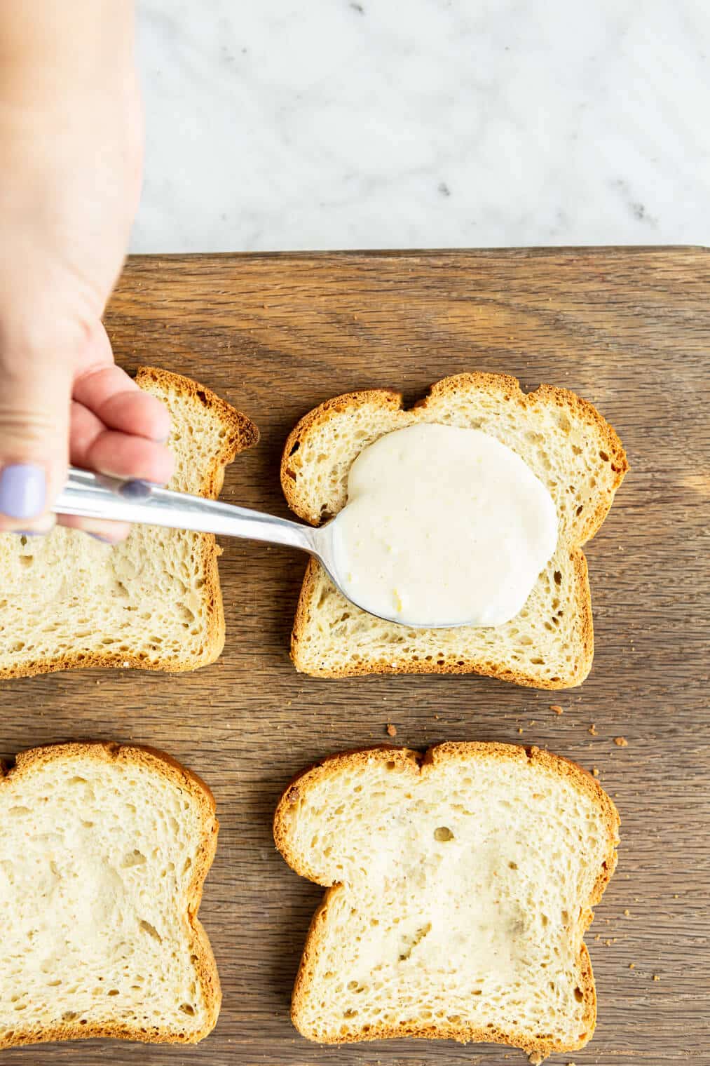 Hand holding spoonful of custard adding it to slice of bread on a wooden cutting board with 4 other slices of bread.