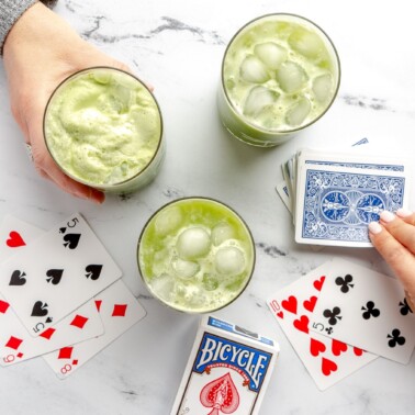 There are three glasses of green matcha iced tea on a white and black marble surface along with a deck of playing cards. There is a hand holding a glass of matcha tea and a hand drawing a card from the top of the deck.