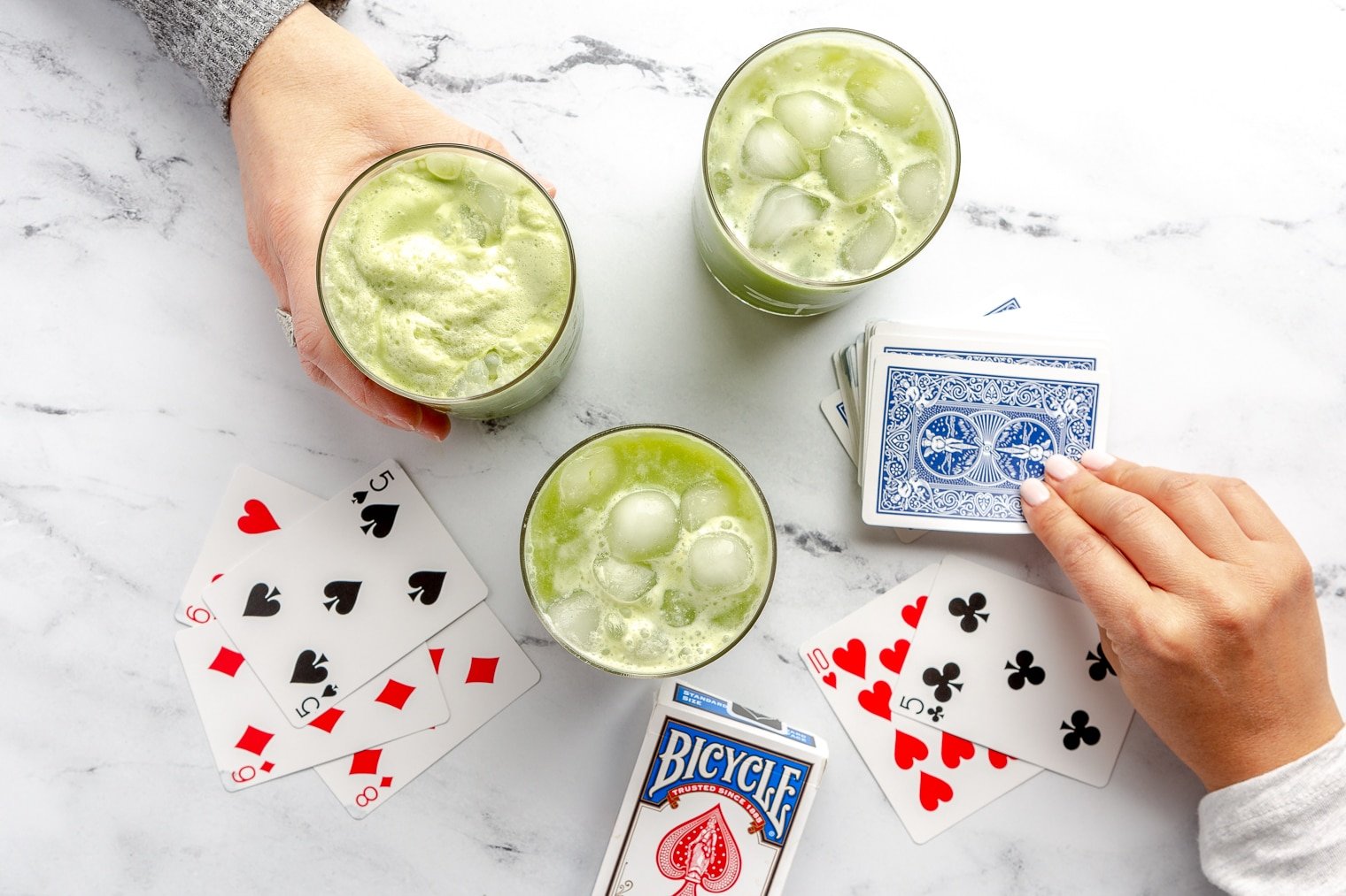 There are three glasses of green matcha iced tea on a white and black marble surface along with a deck of playing cards. There is a hand holding a glass of matcha tea and a hand drawing a card from the top of the deck.