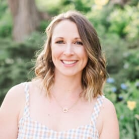 A headshot of Steph Greunke outside, standing in front of greenery and flowers.