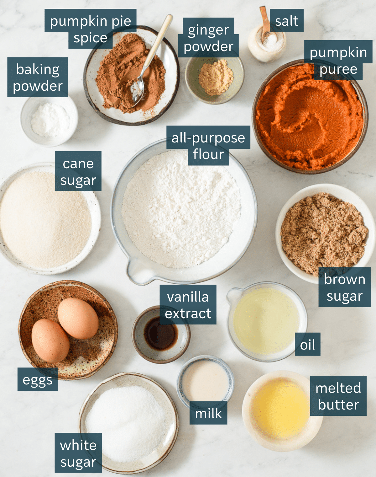 All of the ingredients needed for classic pumpkin bread in different sized bowls and plates on a marble surface.