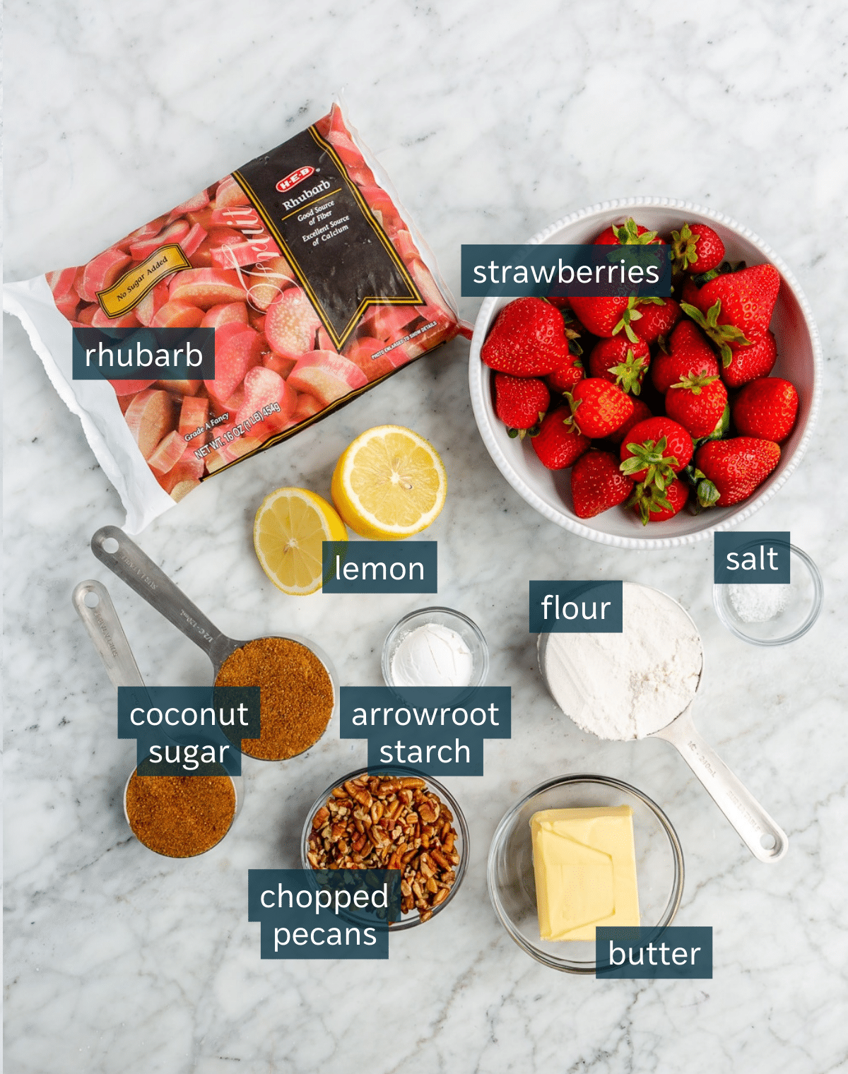 Ingredients for strawberry rhubarb crisp sit in a variety of bowls and measuring cups on a marble countertop.