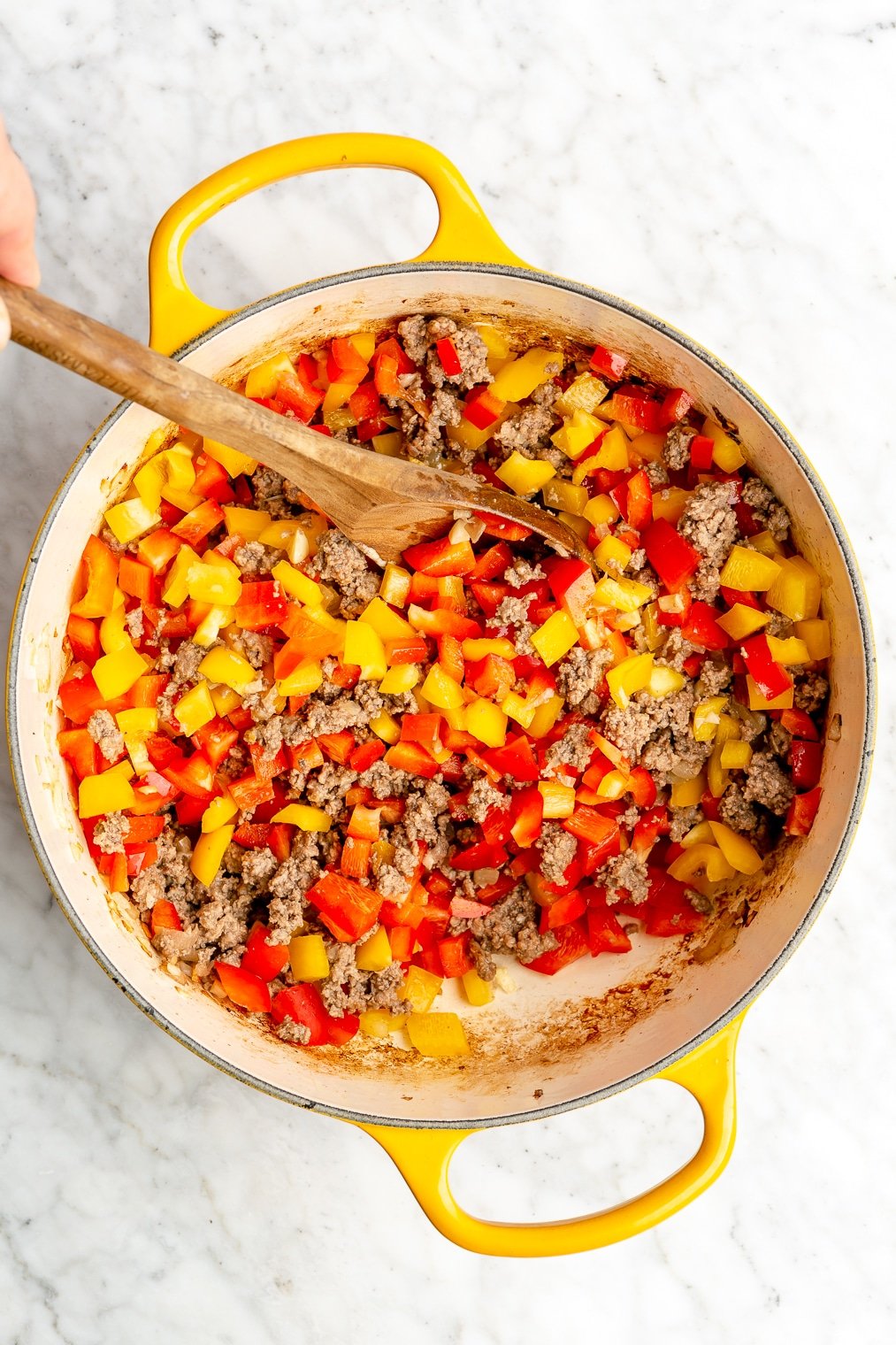A person stirring chopped yellow and red bell peppers into a pot of browned ground beef and sauteed onions and garlic.