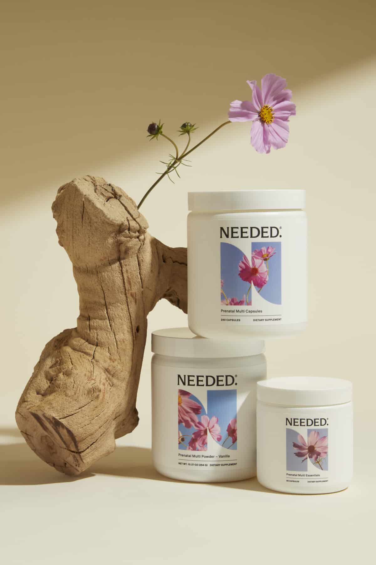 3 Needed nutrition products propped up on a piece of wood with a pink flower on top.