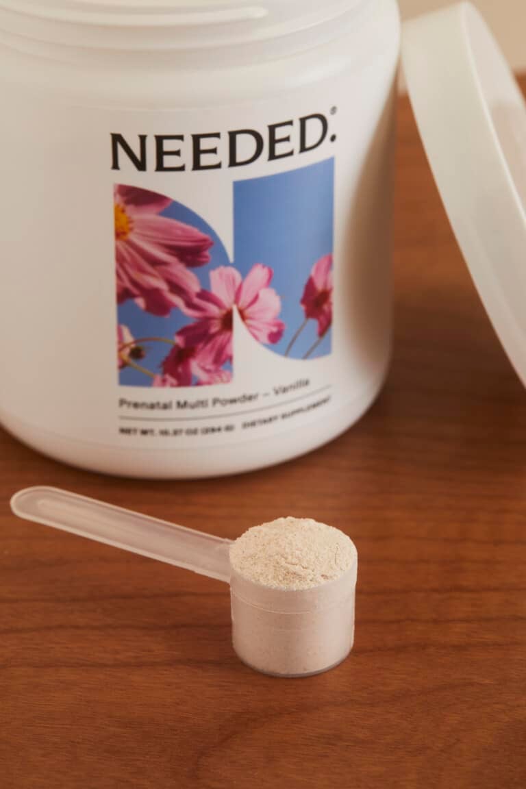 A container of Needed prenatal powder with a scoop in the scooper sitting in front of it on a wooden countertop.