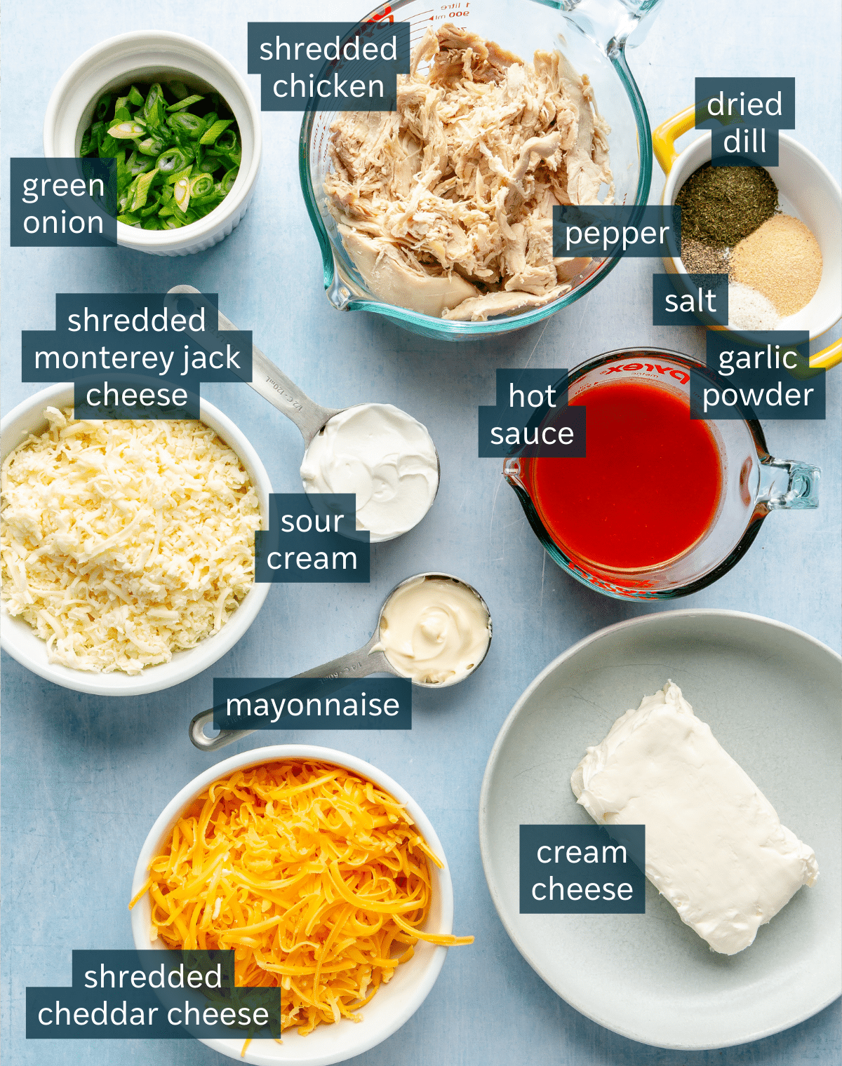 Ingredients for buffalo chicken dip sit in a variety of bowls on a light blue background.