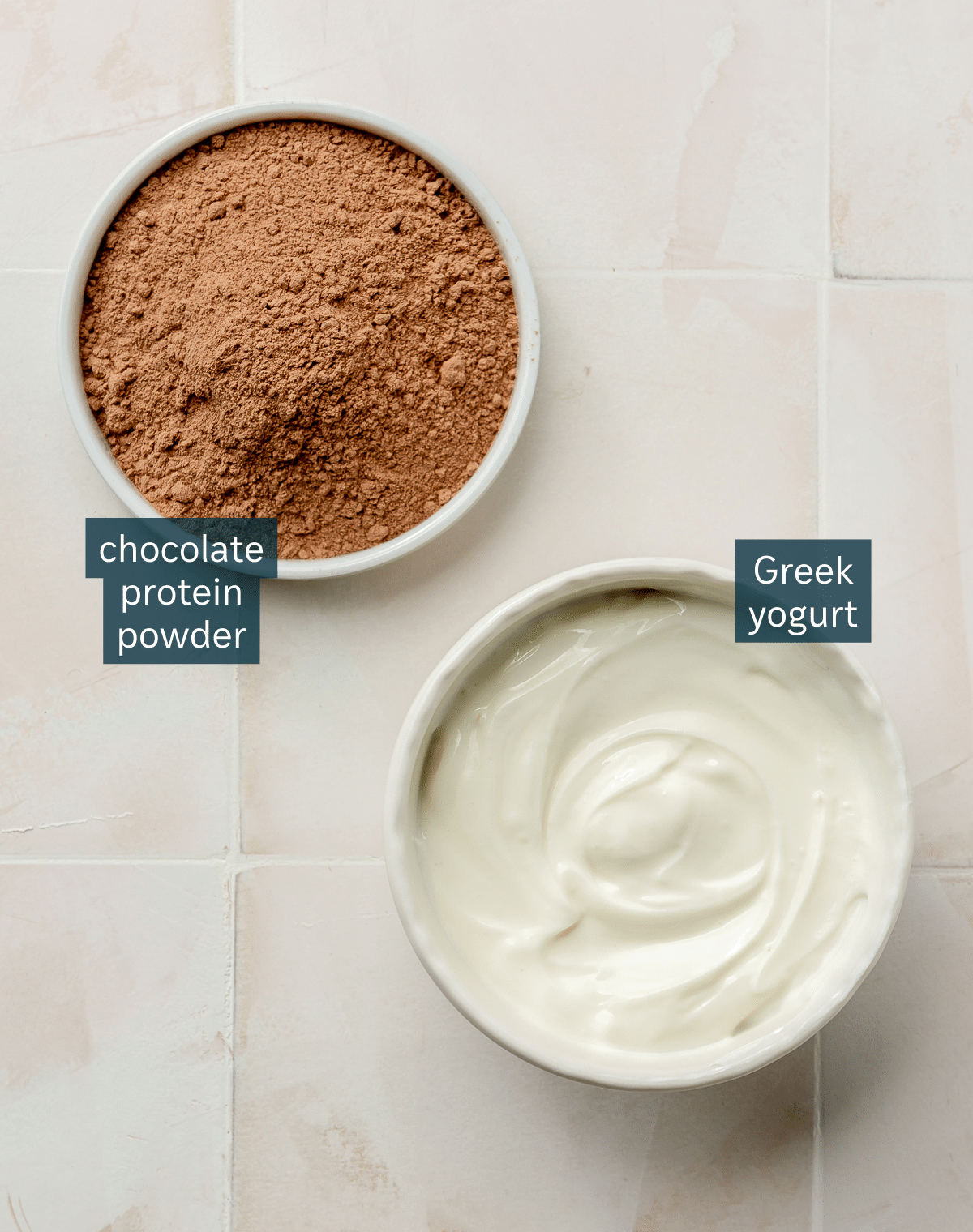 Ingredients for chocolate protein pudding sit in two different bowls on a white tiled countertop.