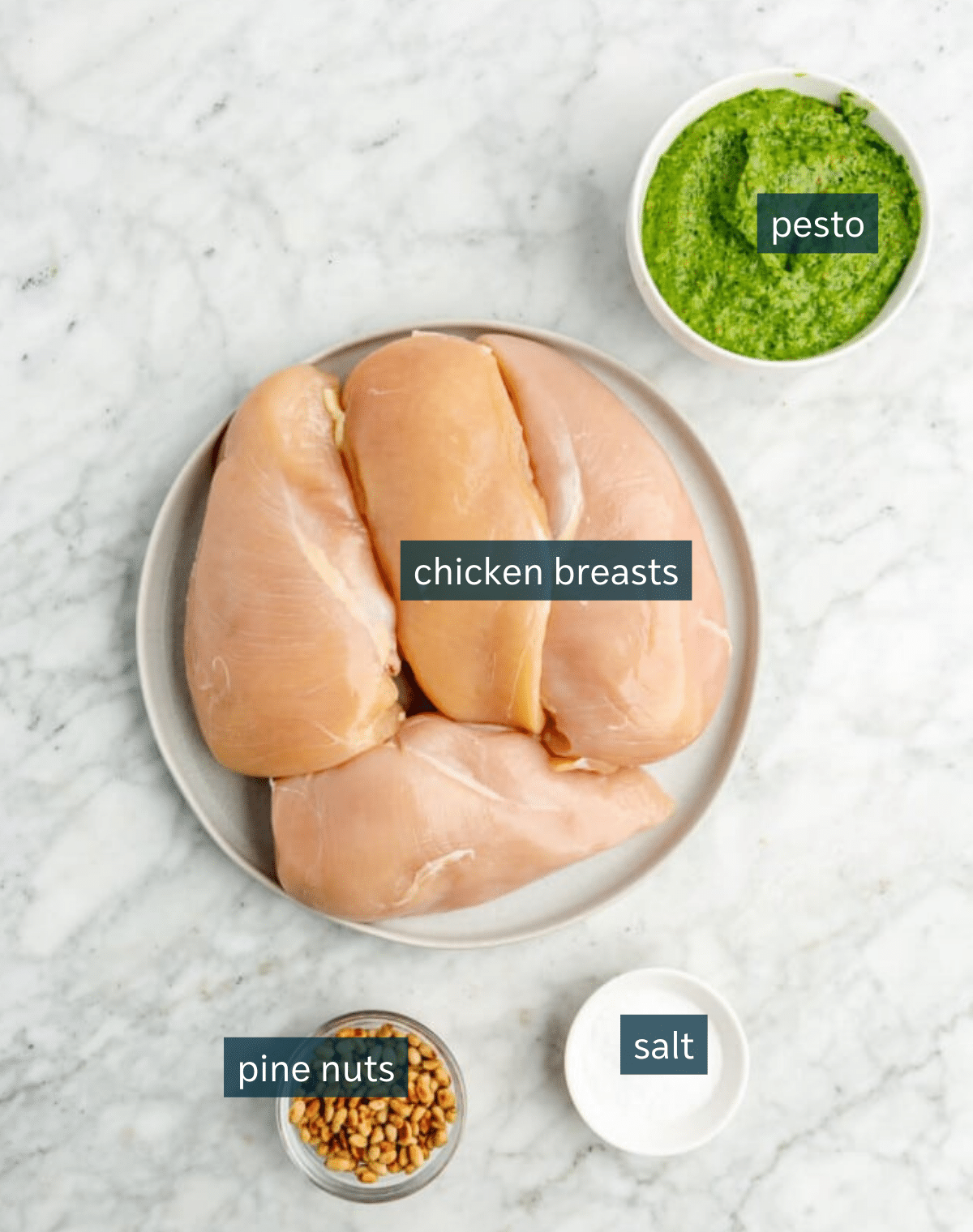Ingredients for pesto chicken salad sit in a variety of bowls on a marble countertop.