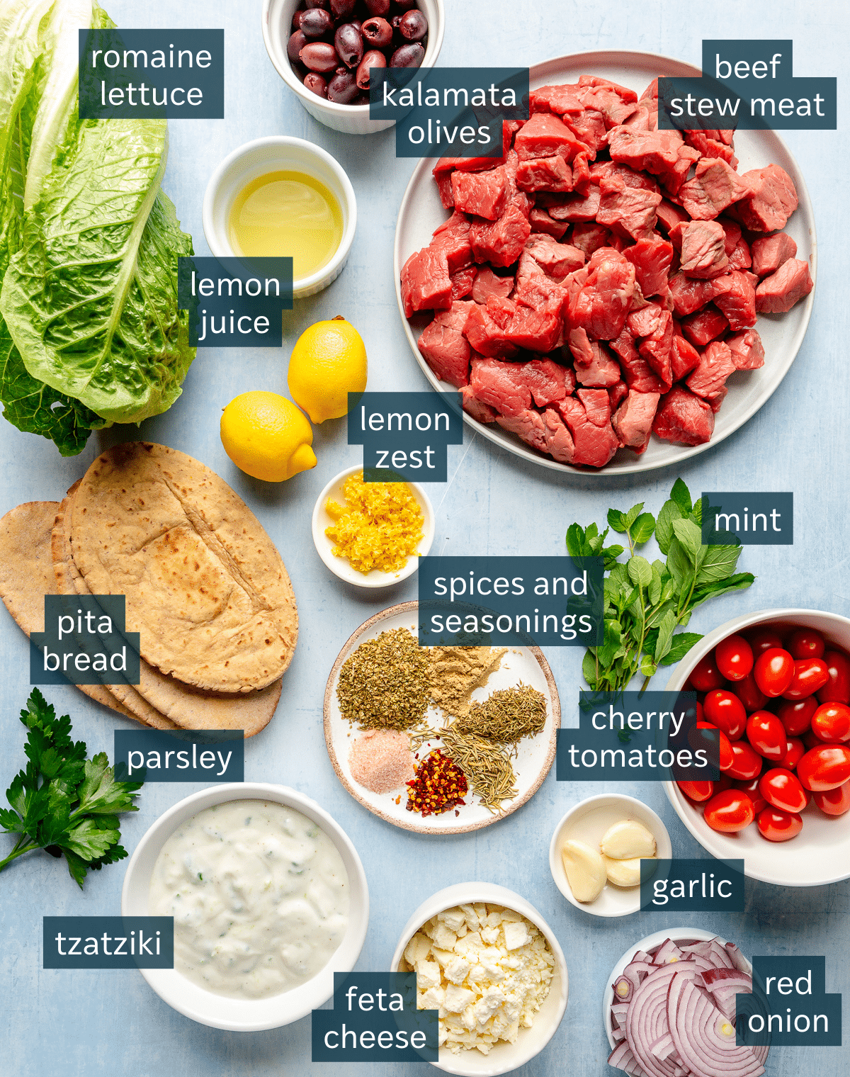 Ingredients for beef gyros sit in a variety of bowls on a light blue surface.