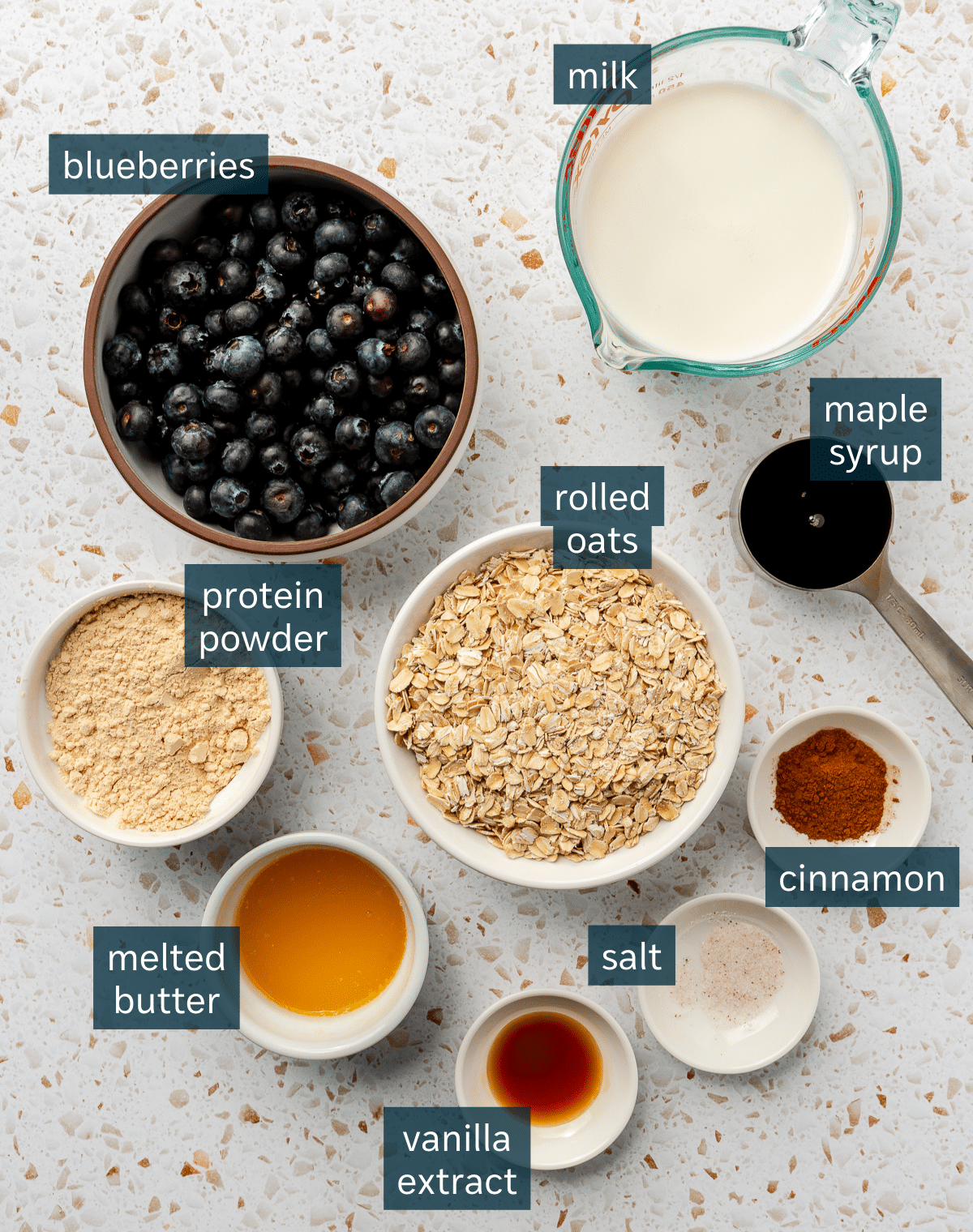 Ingredients for high protein oatmeal bake sit in a variety of bowls on a white speckled countertop.