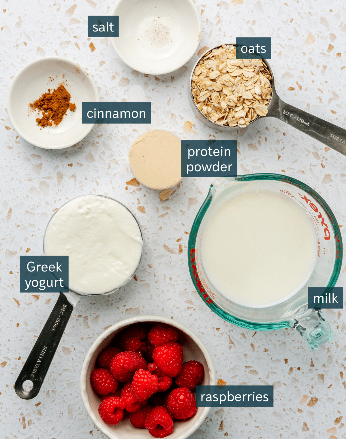 All of the ingredients needed for high protein oatmeal in different sized bowls and plates on a terrazzo surface. 