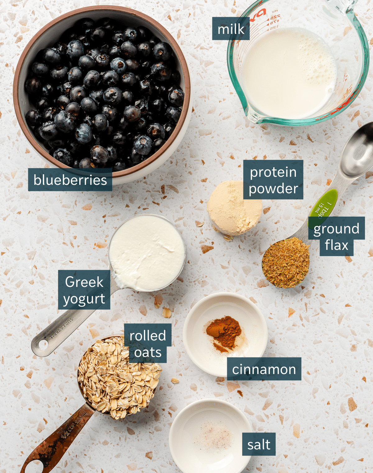 Ingredients for high protein overnight oats sit in a variety of bowls on a white speckled countertop.