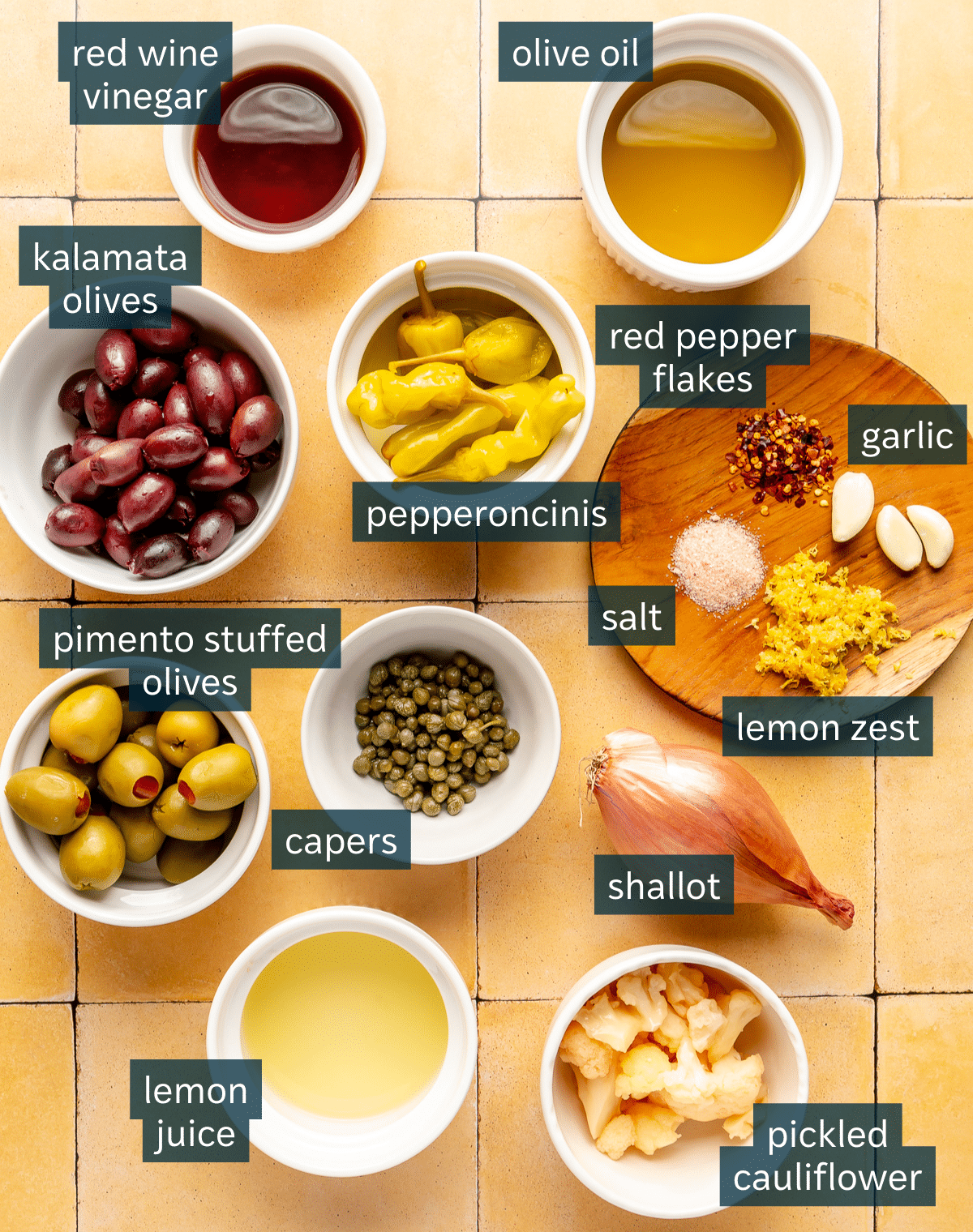 Ingredients for olive tapenade sit in a variety of bowls on a yellow tiled surface.