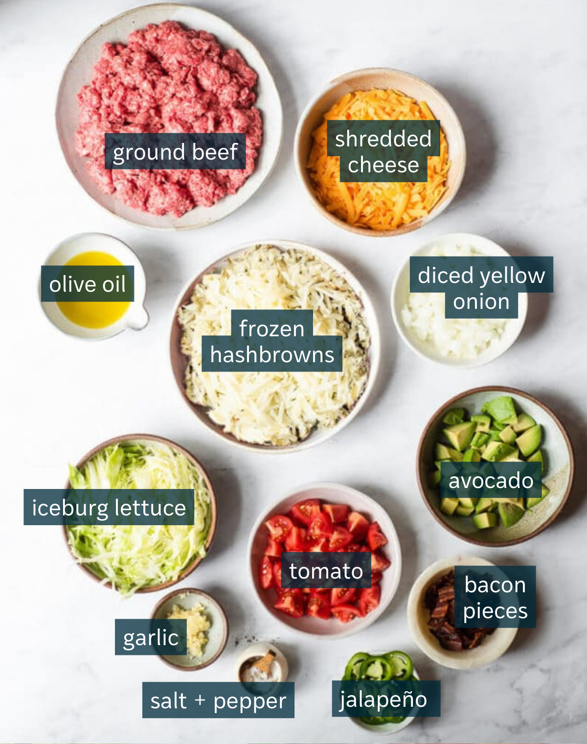 Ingredients for cheeseburger casserole sit in a variety of bowls on a white countertop.