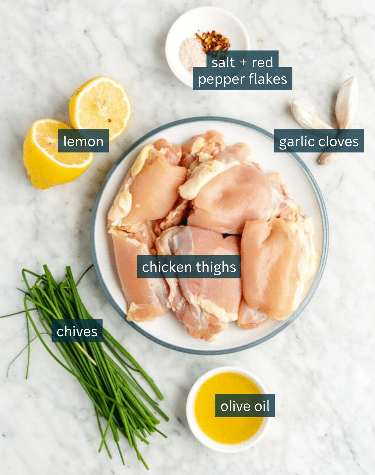 Ingredients for lemon garlic chicken thighs sit in a variety of bowls on a marble surface.