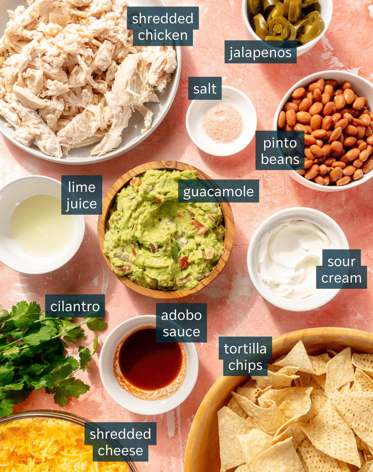 Ingredients for sheet pan nachos sit in a variety of bowls on a light pink surface.