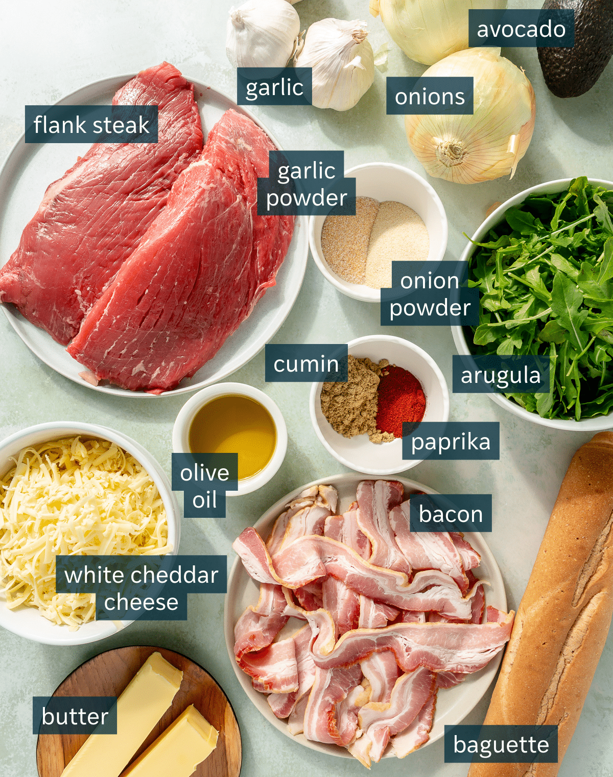 Ingredients for steak sandwiches sit in a variety of bowls on a light blue surface.