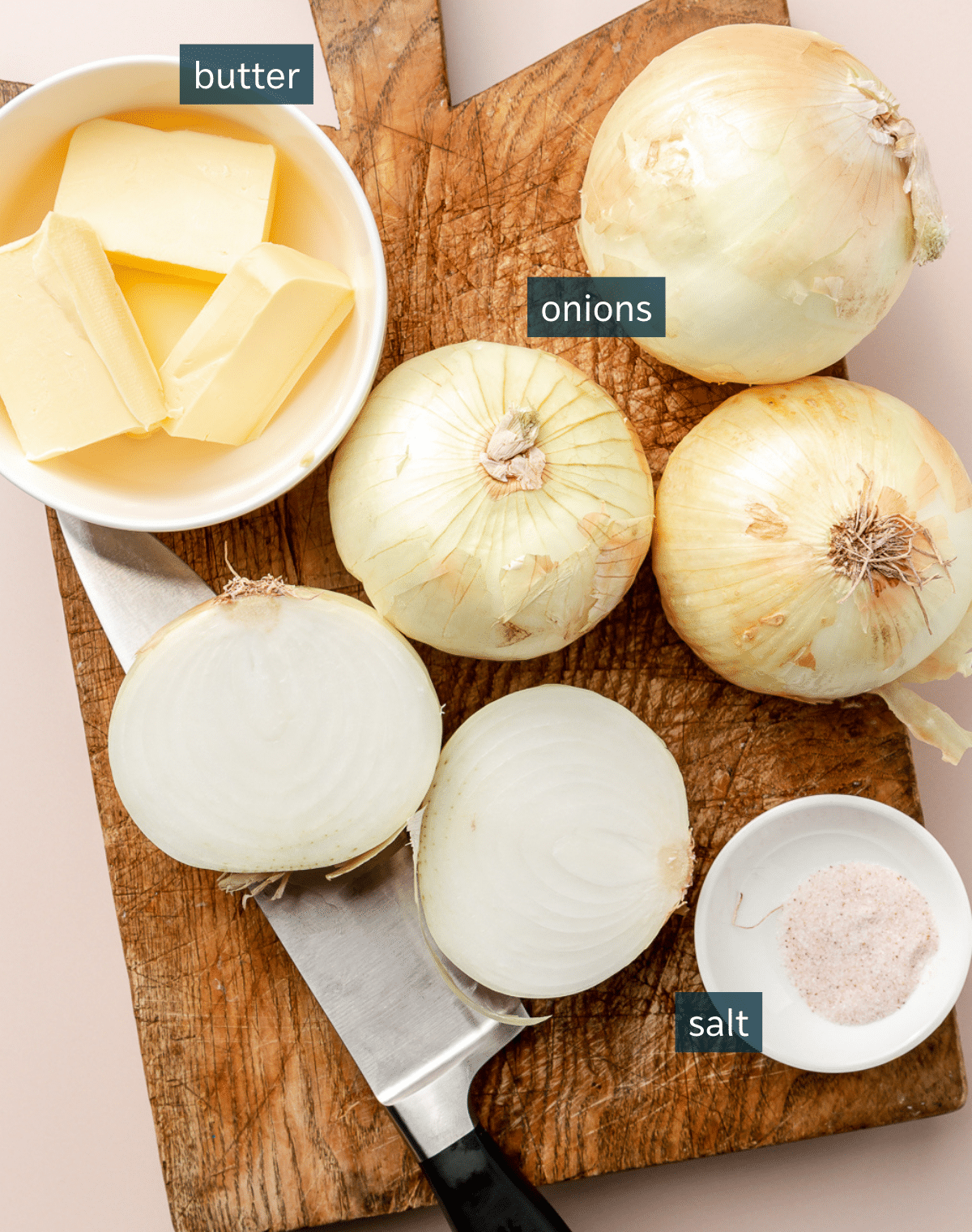 A wooden cutting board with 4 onions (one cut in half), a small bowl of salt, and a bowl of butter.